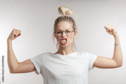 I can everything. Happy young blonde woman in white t-shirt showing biceps on her arms. Emotional portrait with white background of expressive girl with blonde hair bun and spectacles.