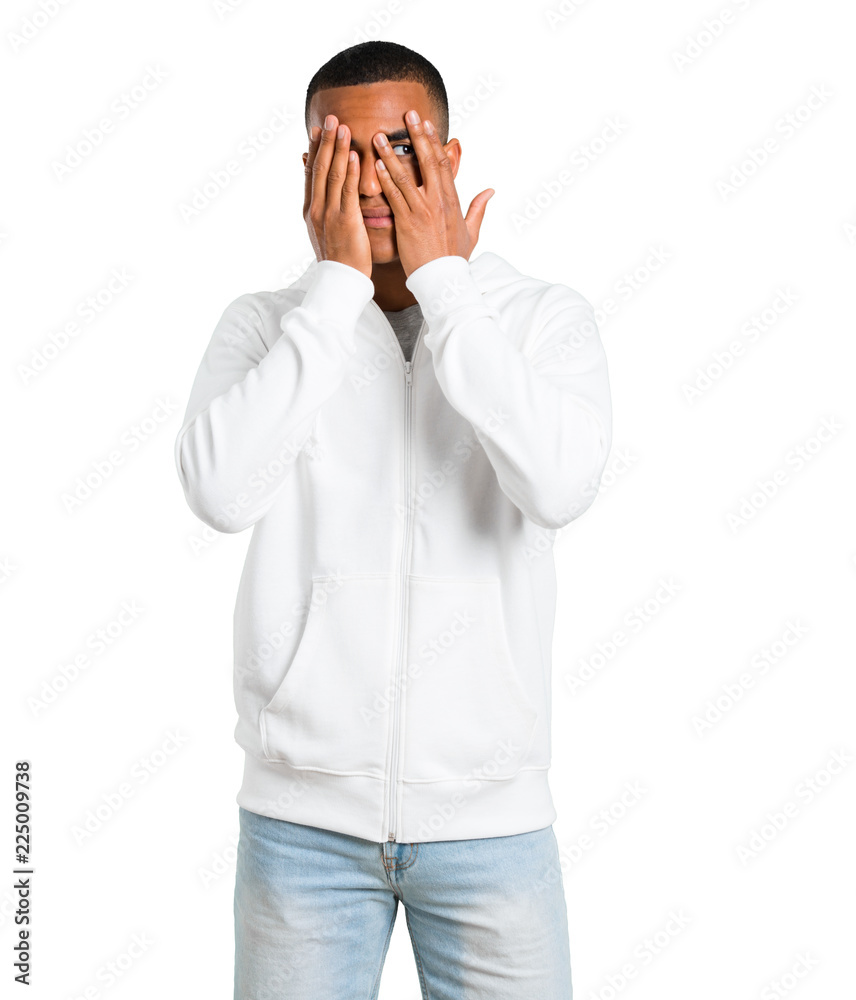 Dark-skinned young man with white sweatshirt covering eyes by hands and looking through the fingers on isolated white background Dark-skinned young man with white sweatshirt covering eyes by hands and looking through the fingers on isolated white background