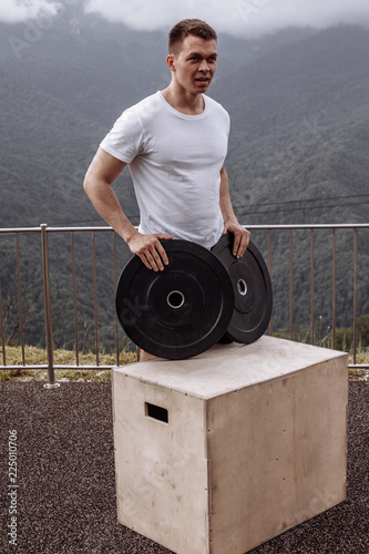 Strong caucasian crossfit male instructor posing with barbell plate over forest and sky background, strong muscles shoulders biceps triceps chest. photo