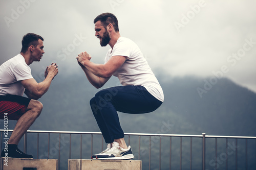 Cross-fit male athletes doing plyometric exercise. Jumping on the box. Phase touchdown, close up. Outdoor workout over mountains and foggy sky background photo