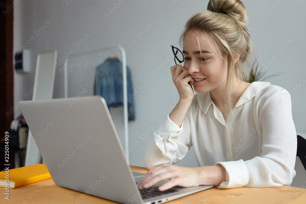 Candid shot of positive young female freelancer with messy hairstyle using generic electronic portable device at wooden desk, working remotely from home office, having absorbed interested look Candid shot of positive young female freelancer with messy hairstyle using generic electronic portable device at wooden desk, working remotely from home office, having absorbed interested look
