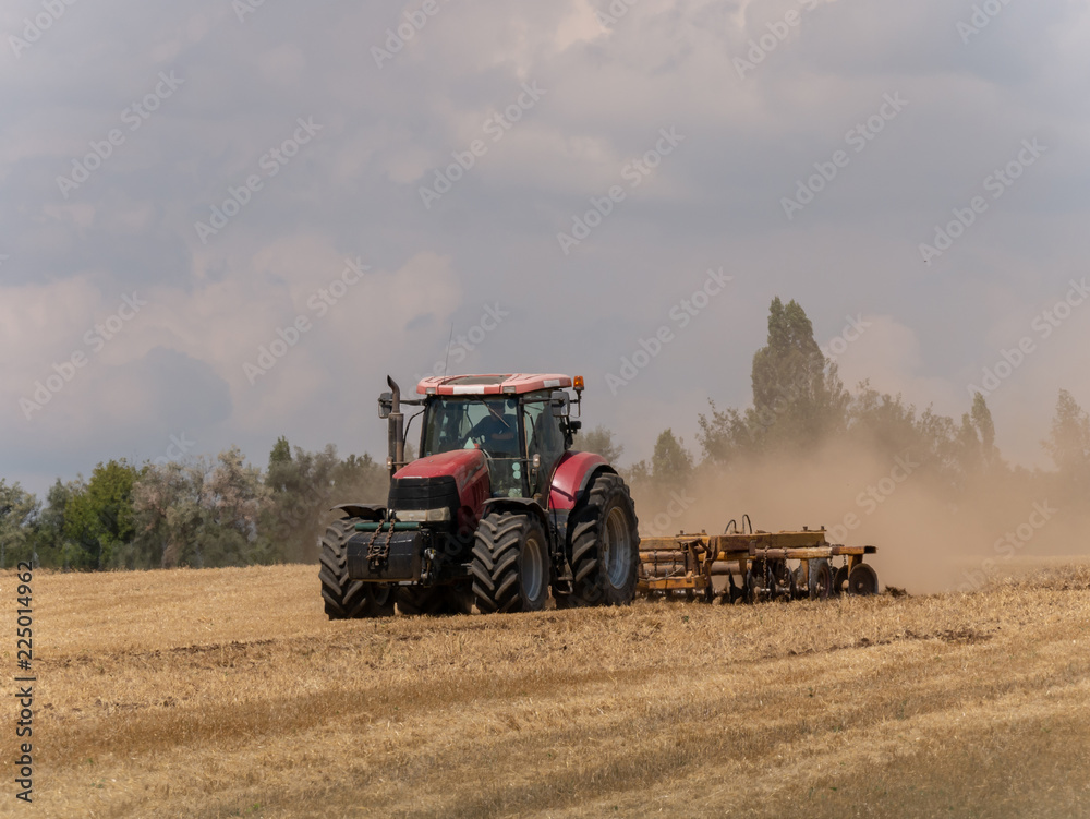 Obraz premium Red tractor with plow on harvest field. Summer field and red tractor. Tractor on harvest field. Lowing after harvest. Plowing on the field.