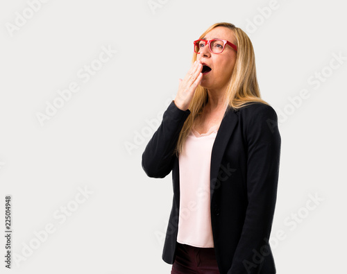 Middle-age blonde business woman with black jacket yawning and covering wide open mouth with hand. Sleepy expression on isolated grey background