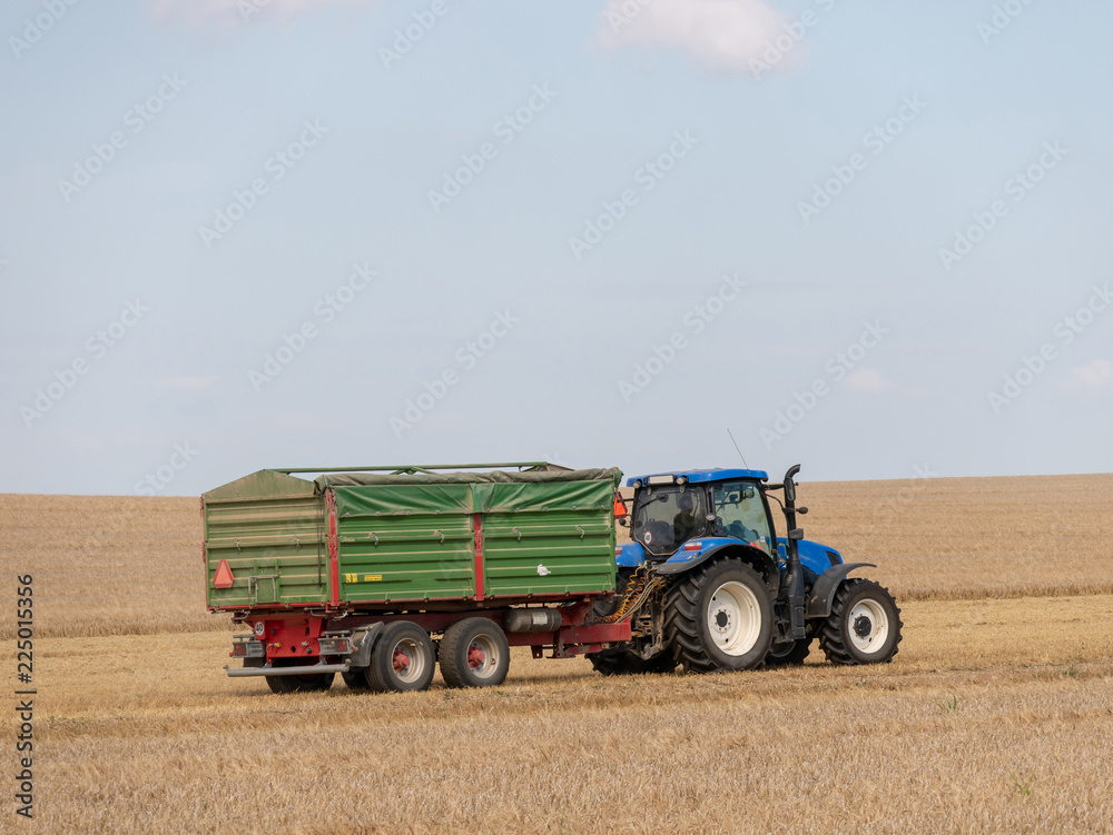 Obraz premium Blue tractor with trailer on the harvest field. Summer field and blue tractor with trailer. Harvest field, blue sky and tractor.