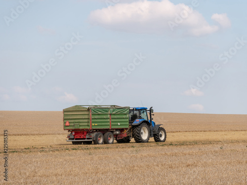 Blue tractor with trailer on the harvest field. Summer field and blue tractor with trailer. Harvest field  blue sky and tractor.