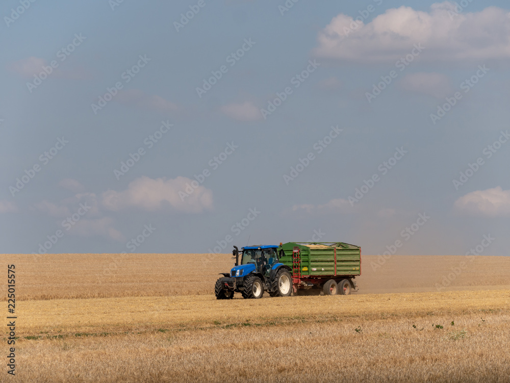 Blue tractor with trailer on the harvest field. Summer field and blue tractor with trailer. Harvest field, blue sky and tractor. Blue tractor with trailer on the harvest field. Summer field and blue tractor with trailer. Harvest field, blue sky and tractor.