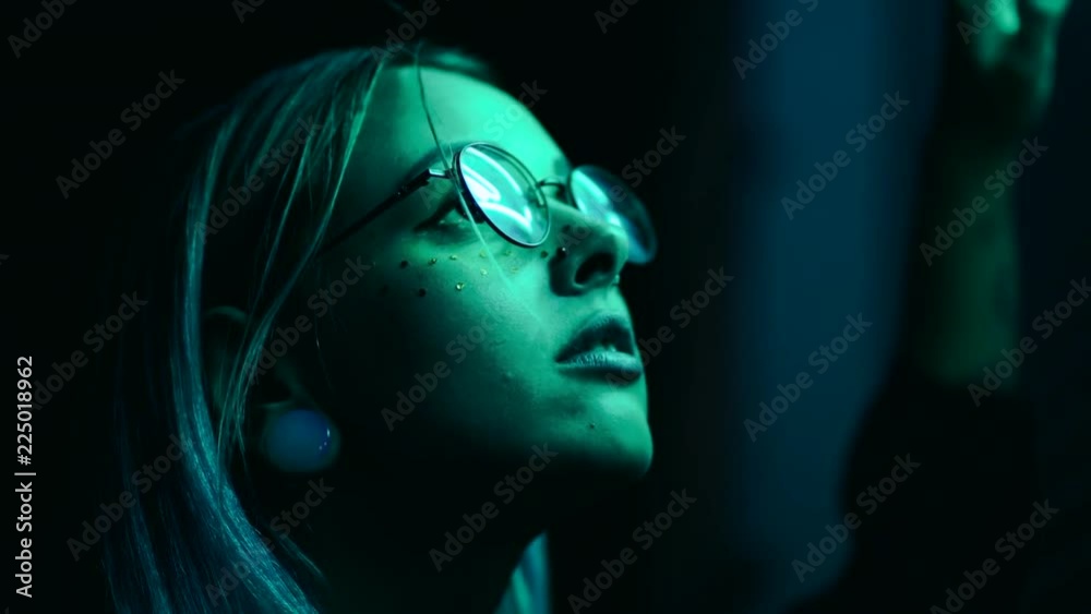 Millennial enigmatic pretty girl with unusual dyed hairstyle near glowing neon wall at night. Blue hair, golden sequins as freckles,nose piercing. Mysterious hipster teenager in glasses.