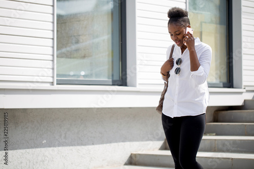 Happy african-american woman in white classy shirt looking at camera while sitting on staicase outdoors. Technology, communication and big city lifestyle concept, copy space photo