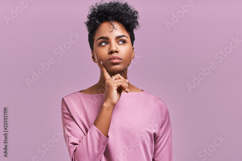 Pensive attractive curly African American female being deep in thoughts, raises eye, wears fashionable clothes, stands against lavender wall