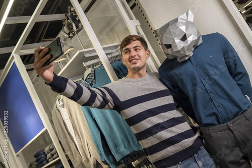 Selfie while shopping. A guy in a striped sweater in a store with clothes takes pictures of himself on the phone next to mannequins with an animal head.