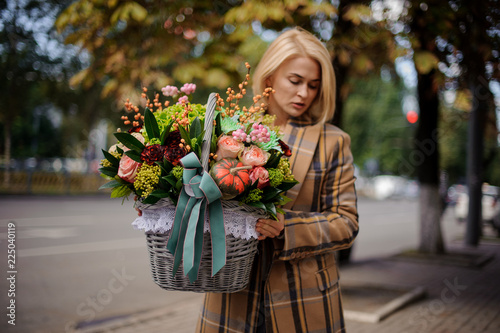 Young blonde woman holding a big wicker basket of flowers against the city