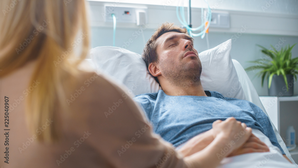 In the Hospital Sick Man Lying on the Bed, His Visiting Wife Hopefully Sits Beside Him Holds His Hands and Hopes for Recovery. In the Hospital Sick Man Lying on the Bed, His Visiting Wife Hopefully Sits Beside Him Holds His Hands and Hopes for Recovery.