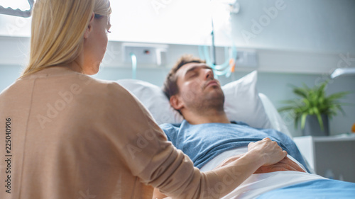 In the Hospital Sick Man Lying on the Bed, His Visiting Wife Hopefully Sits Beside Him Holds His Hands and Hopes for Recovery.