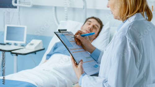 Friendly Female Doctor Visits Smiling Recovering Man who is Lying in Bed, She Asks Him Questions and Fills Medical Chart.