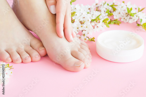 Young woman's hand touching her perfect feet. White jar of natural herbal cream on pastel pink background. Care about clean, soft and smooth skin. Beautiful branch of cherry blossoms. Fresh flowers.