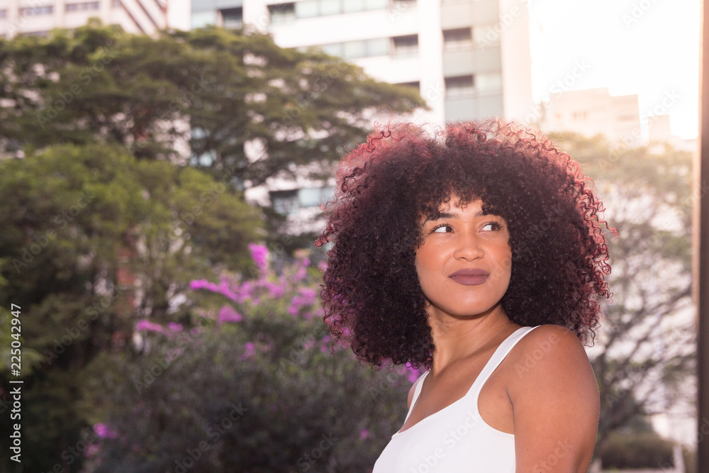 Portrait of woman looking to the side and smiling. In the city during summer, builindgs and tree tops. She is black, on her early twenties, Afro style frizzy hair.
