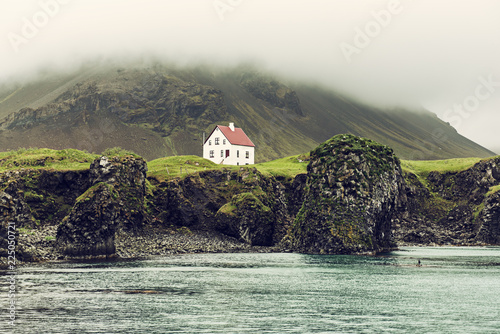 Платно Lonely icelandic house with red roof on the sea coast with green grass meadow, rocks anf foggy sky