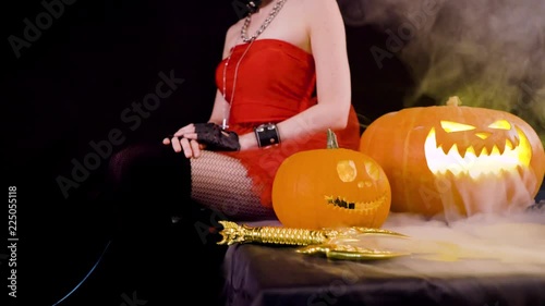 Angry evil devil girl with horns grins, want to kill, murder, leads thumb along the neck, wicked smile. Celebrate halloween with scary pumpkins on dark black background in smoke