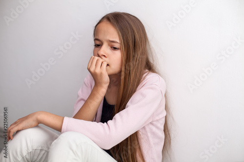 Worried teenage girl biting her nails sitting on the floor by the wall