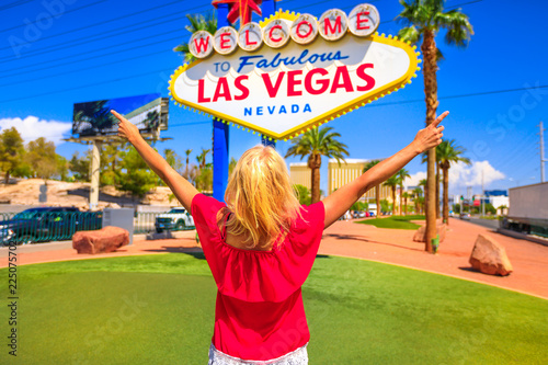 Carefree woman with raised arms at Las Vegas Nevada Sign on blurred background, popular landmark on Las Vegas Strip at entrance of the city. Happy tourist in Nevada, Unites States. Blue sky.