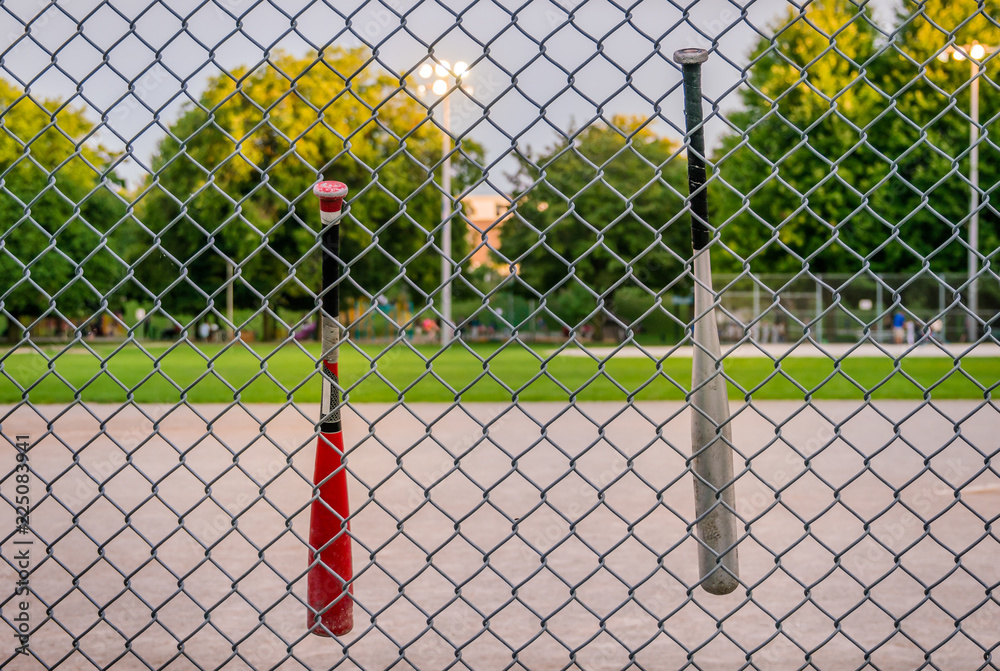 Obraz premium Baseball bats hanging from a wired fence in front of a blurred basedball field, at twilight,