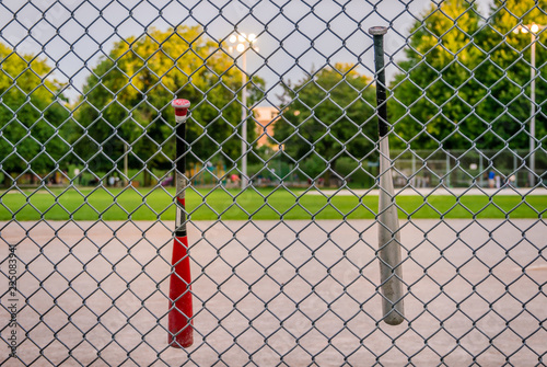 Baseball bats hanging from a wired fence in front of a blurred basedball field, at twilight,
