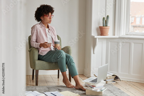 Thoughtful successful female startupper enjoys cappuccino beverage, holds takeaway cup, sits in armchair, has break after work, uses cell phone and laptop computer for searching indormation online. photo