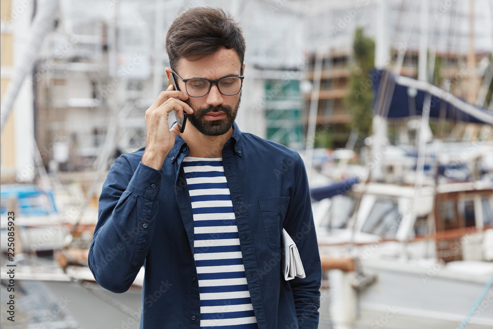 Horizontal shot of thoughtful guy makes call to customer support, checks his account balance, wears fashionable outfit, waits for someone in harbour, enjoys communication. Lifestyle concept. Horizontal shot of thoughtful guy makes call to customer support, checks his account balance, wears fashionable outfit, waits for someone in harbour, enjoys communication. Lifestyle concept.
