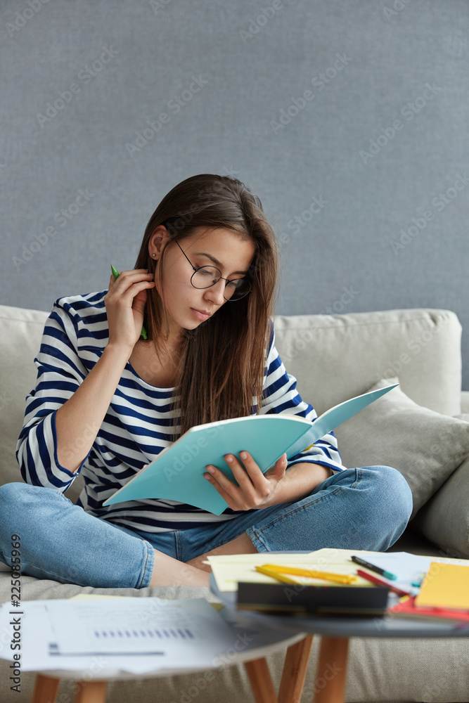Pretty student with dark hair focused in textbook, prepares for writing course paper or upcoming exam, sits crossed legs on comfortable sofa against modern apartment interior. Studying concept