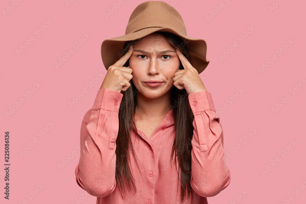 Frustrated woman with displeased facial expression, has bad memory, keeps both index fingers on temples, tries concentrate on important task, look directly into camera, models indoor against pink wall