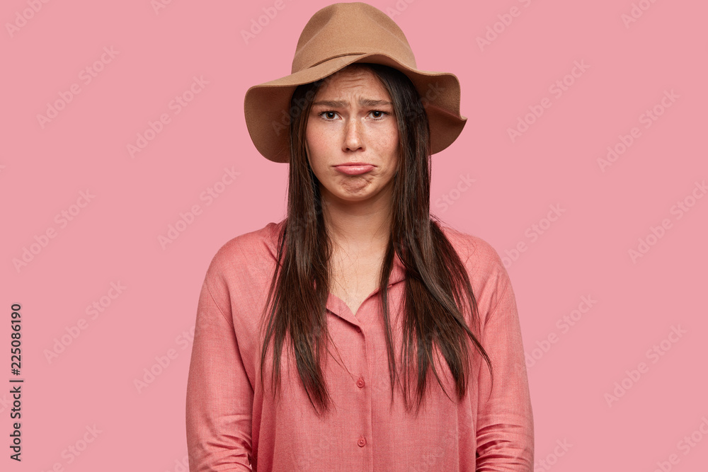 Indoor shot of dissatisfied woman has placid discontent facial expression, purses lips in displeasure, being in low spirit, dressed in fashionable hat, poses against pink background. Negative feeling Indoor shot of dissatisfied woman has placid discontent facial expression, purses lips in displeasure, being in low spirit, dressed in fashionable hat, poses against pink background. Negative feeling