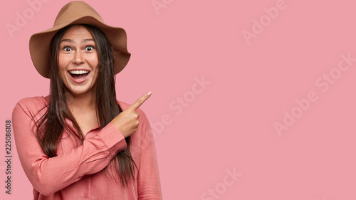 Horizontal shot of pretty of young freckled European woman points with index finger at copy space, being amazed by unusual item, dressed in fashionable pink shirt in one tone with background.