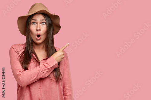 Surprised brunette Caucasian woman with astonished expression, keeps mouth opened from wonder, shows with index finger at blank space, advertises unusual item, isolated over pink background.