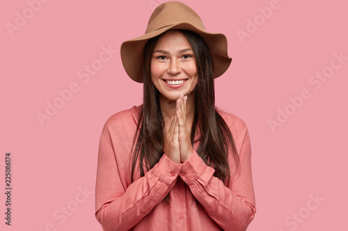Horizontal shot of smiling beautiful woman with satisfied expression, keeps hands together from joy, hears positive news from interlocutor, wears stylish shirt and headgear isolated on pink background