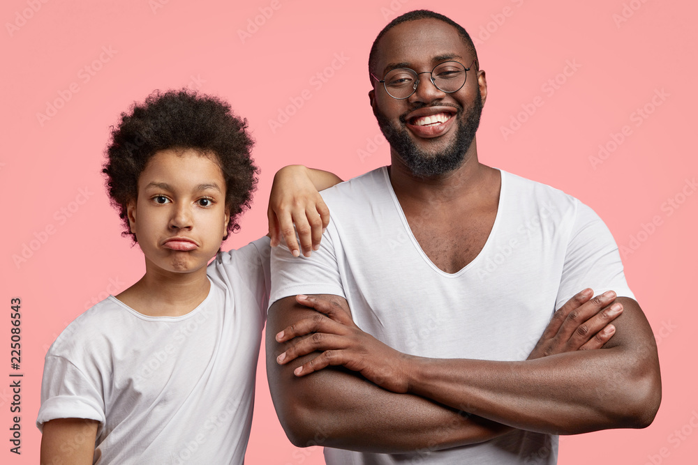 Photo of smiling satisfied black male adult has shining smile, keeps arms folded, spends free time with son, going to have shopping together, pose aganist pink background. Generation concept Photo of smiling satisfied black male adult has shining smile, keeps arms folded, spends free time with son, going to have shopping together, pose aganist pink background. Generation concept