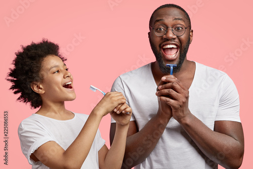 Happy dark skinned joyful dad and son hold toothbrsuh and shaver, have morning routine together, clean teeth and shave beard in bathroom, have hygienic procedures, isolated over pink background.