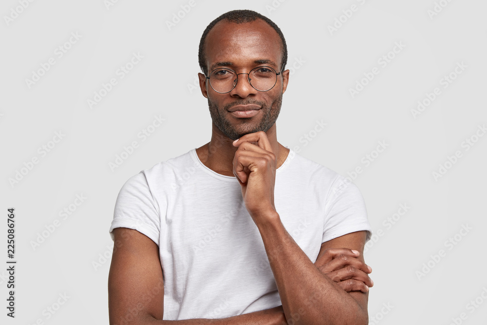 Studio shot of serious black man keeps hand under chin, wears transparet  glasses and casual t shirt, keeps hands partly crossed, poses against white  background, thinks about something, being clever Stock Photo