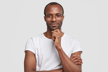 Studio shot of serious black man keeps hand under chin, wears transparet glasses and casual t shirt, keeps hands partly crossed, poses against white background, thinks about something, being clever