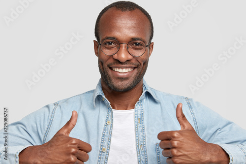 I agree with your opinion. Photo of glad dark skinned adult man with stubble raises thumbs and smiles broadly, approves interesting idea, wears transparent glasses, poses against white background photo