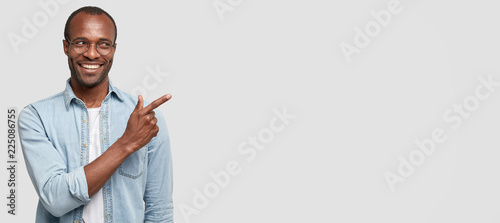 Medium length shot of attractive black man smiles brightfully, dressed in fashionable denim shirt, points at white wall with index finger, advertises something not expensive and of good quality photo