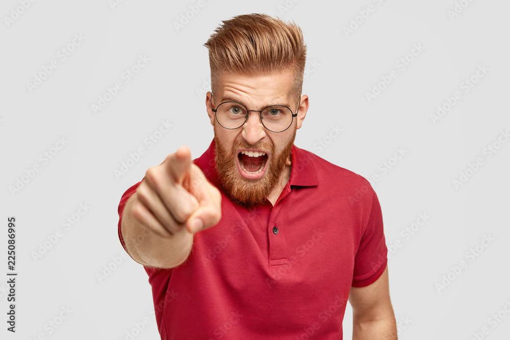 Portrait of peevish bearded guy with ginger trendy hairstyle, yells angrily at somebody, points with index finger directly at camera, dressed in red t shirt, blames you in doing wrong things Portrait of peevish bearded guy with ginger trendy hairstyle, yells angrily at somebody, points with index finger directly at camera, dressed in red t shirt, blames you in doing wrong things