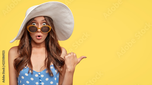 Stunned beautiful young European woman with dark wavy hair, scared expression, dressed in fashionable outfit, ready for summer vacation, indicates aside at blank copy space, yellow background