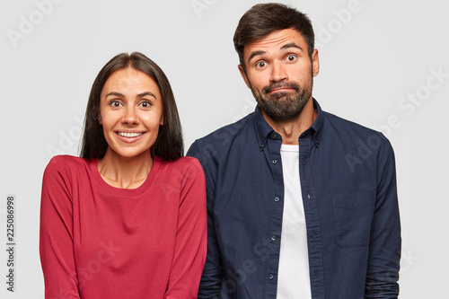 Puzzled uncertain young woman and man look with doubtful expressions at camera, try to make decision, stand against white background. Girlfriend, boyfriend express apathy ask: So what do now?