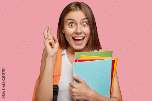 Photo of cheerful brunette woman with overjoyed look keeps fingers crossed, believes in everything good, holds books, has positive expression, poses against pink background. Body language concept