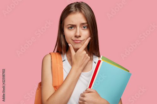 Pensive attractive Cauasian girlfriend holds chin and looks with puzzlement, has straight hair, carries rucksack with school object, holds textbooks, poses against pink background. Studying concept