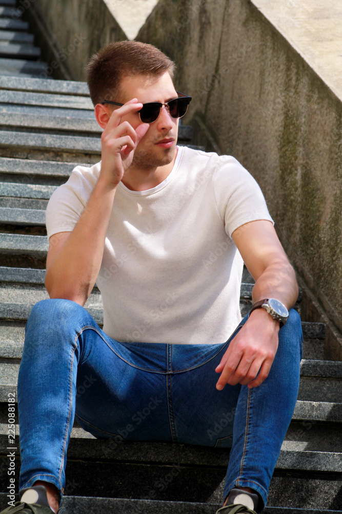 Man sitting alone on steps. Handsome boy with sunglasses. Male model posing for shooting, sitting on old stairs. Portrait of cool guy sitting on marble staircase waiting for someone or something. Man sitting alone on steps. Handsome boy with sunglasses. Male model posing for shooting, sitting on old stairs. Portrait of cool guy sitting on marble staircase waiting for someone or something.