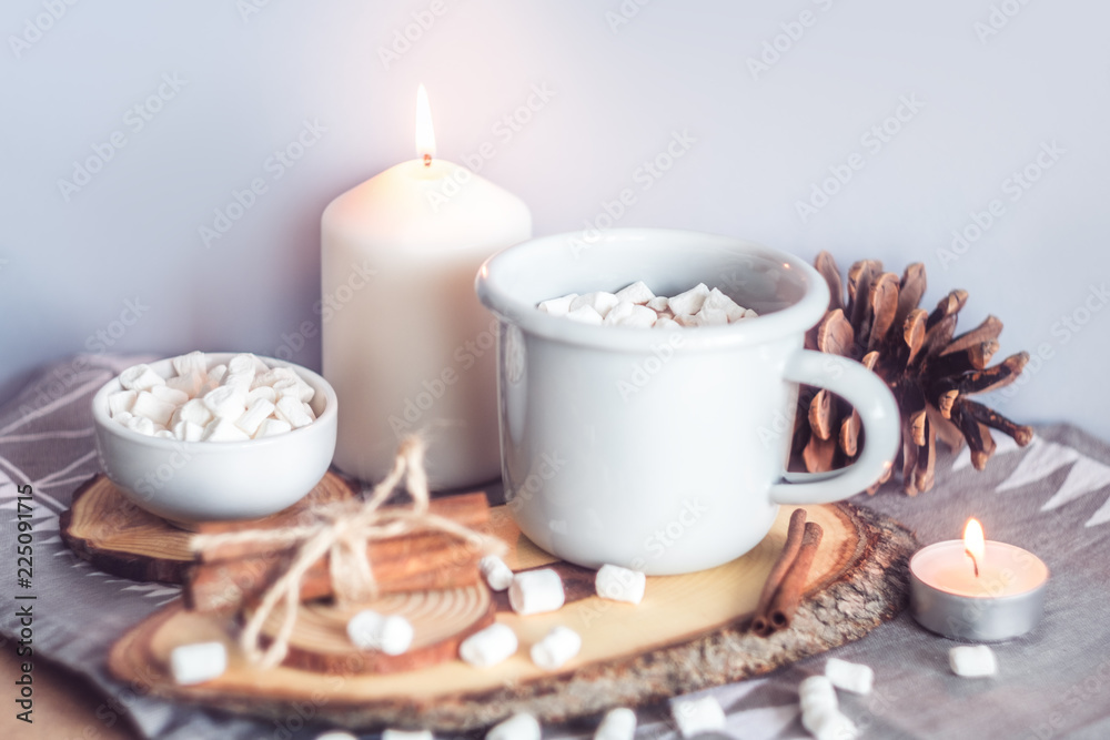 A cup of hot cocoa with marshmallows and candles on a table surrounded by winter or Christmas decor, closeup
