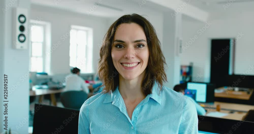 DOLLY IN Portrait of young adult mid 30s beautiful Caucasian female smiling and looking into camera, busy office in the background. 4K UHD 60 FPS SLOW MOTION