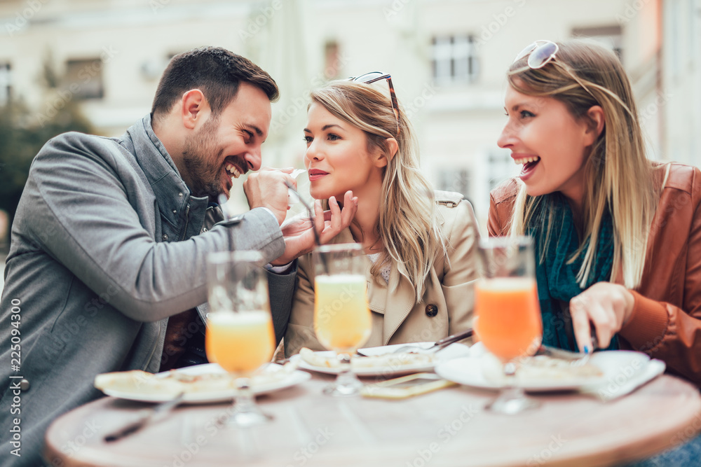 Group of three friends using phone in outdoor cafe on sunny day, eating pizza