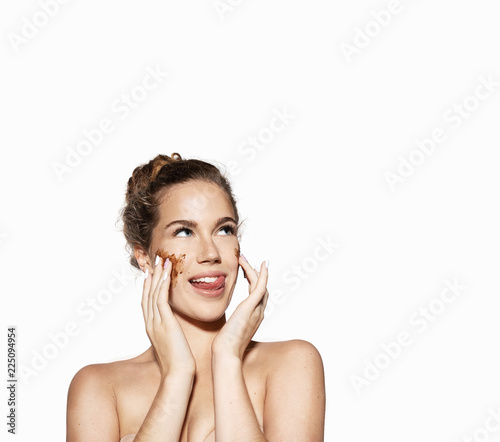 Portrait of a young woman applying a coffee scrub to the face on white background. Pretty girl showing tongue and posing with naked top. Beauty skincare concept.